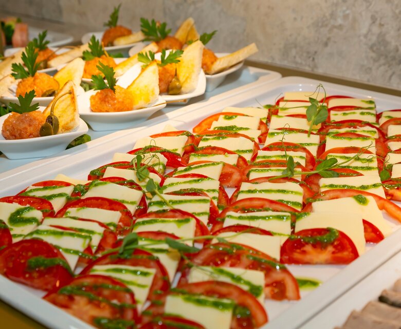 Ein Buffet mit verschiedenen Speisen, darunter eine Platte mit Tomatenscheiben und Käse, garniert mit frischem Basilikum. Im Hintergrund sind kleine Häppchen und gefüllte Teigtaschen zu sehen, die appetitlich angerichtet sind.