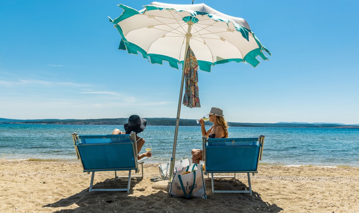 Zwei Frauen sitzen unter einem großen Sonnenschirm am Strand, während sie den Blick auf das ruhige Wasser genießen. Der Sand ist hell und die Atmosphäre ist entspannt und sonnig.