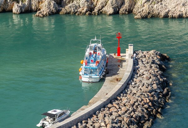 Ein Hafen mit zwei Booten, eines am Steg und das andere im Wasser. Der Steg ist von Steinen umgeben und führt zu einer kleinen roten Laterne. Im Hintergrund sind grüne Hügel und Felsen zu sehen, die das ruhige Wasser umgeben.