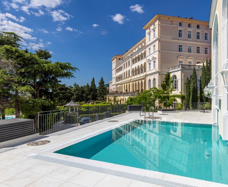 Ein eleganter Außenbereich mit einem klaren Pool, umgeben von grünen Bäumen und einer historischen Hotelarchitektur im Hintergrund. Der Himmel ist blau mit wenigen Wolken, was eine einladende Atmosphäre schafft.