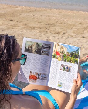 Eine Person in einem blauen Badeanzug sitzt am Strand und liest eine Broschüre über Hotels. Im Hintergrund ist der Sandstrand und das Wasser sichtbar, während die Sonne scheint.