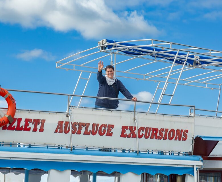 Ein Mann winkt von der oberen Plattform eines Ausflugsschiffs, umgeben von blauem Himmel und weißen Wolken. An der Seite des Schiffs ist ein Rettungsring sichtbar.