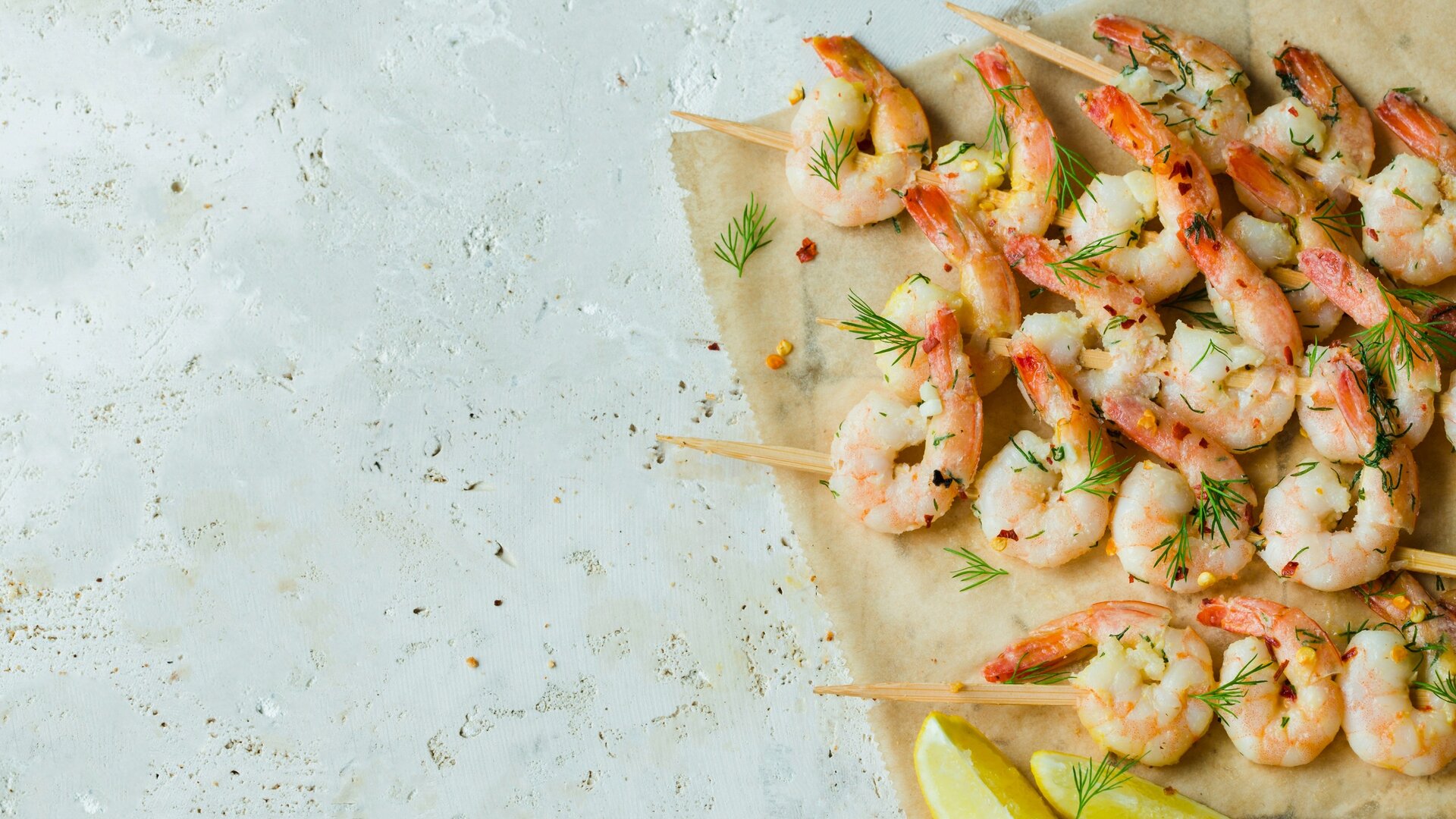 Gegrillte Garnelen auf Holzspießen, garniert mit frischen Kräutern und Zitrone, auf einem Stück Backpapier. Die Garnelen sind saftig und appetitlich angerichtet, umgeben von einer hellen, neutralen Oberfläche.