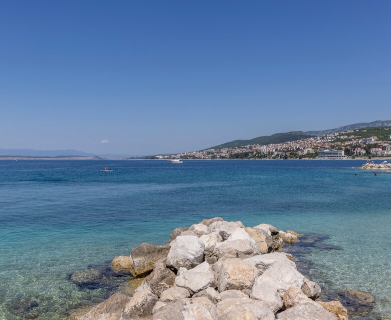 Ein klarer blauer Himmel über einem ruhigen, türkisfarbenen Wasser. Im Vordergrund liegt eine kleine Steininsel, während im Hintergrund eine Küstenstadt mit grünen Hügeln zu sehen ist. Die Atmosphäre ist friedlich und einladend.
