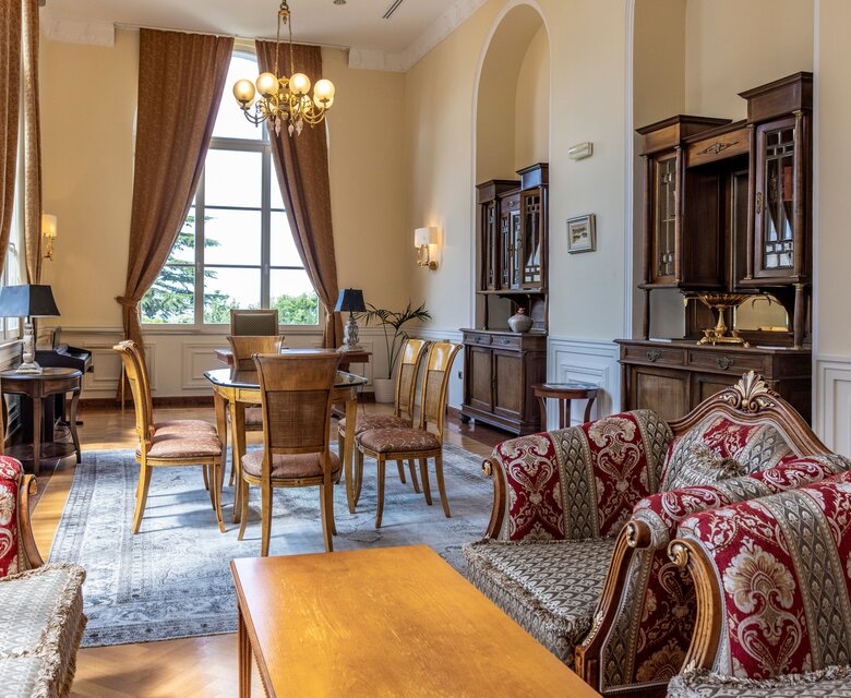 Elegantes Wohnzimmer mit einem großen Tisch und Stühlen, umgeben von stilvollen Möbeln. Große Fenster lassen viel Licht herein, während dekorative Vorhänge und ein Kronleuchter eine gemütliche Atmosphäre schaffen. Die Einrichtung kombiniert klassische und moderne Elemente.
