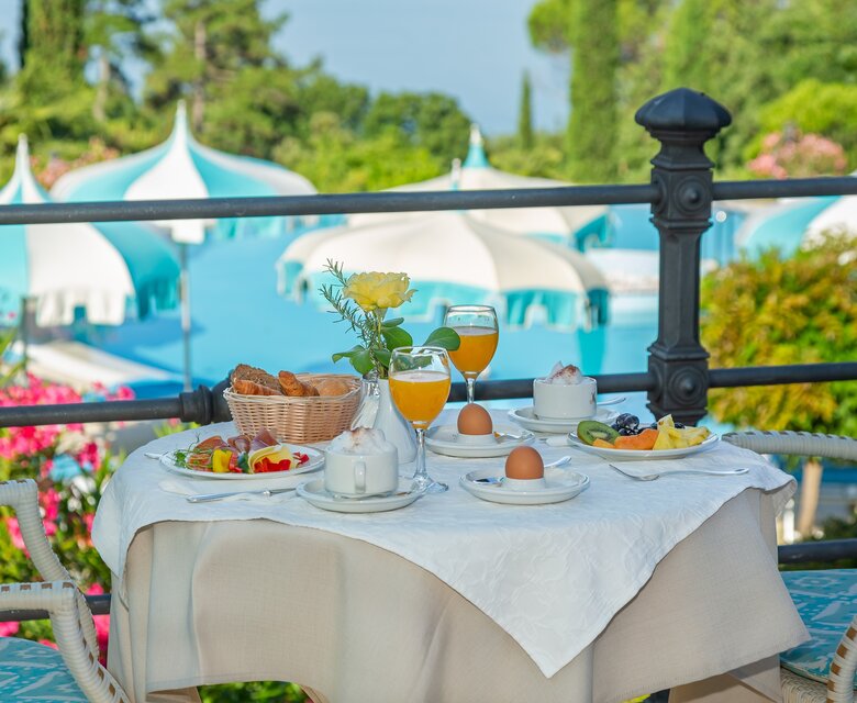 Ein gedeckter Tisch mit Frühstücksgerichten, darunter frisches Obst, Eier und Gebäck, steht auf einer Terrasse. Im Hintergrund sind blühende Pflanzen und ein blauer Pool mit weißen Sonnenschirmen zu sehen, was eine entspannte Urlaubsatmosphäre schafft.