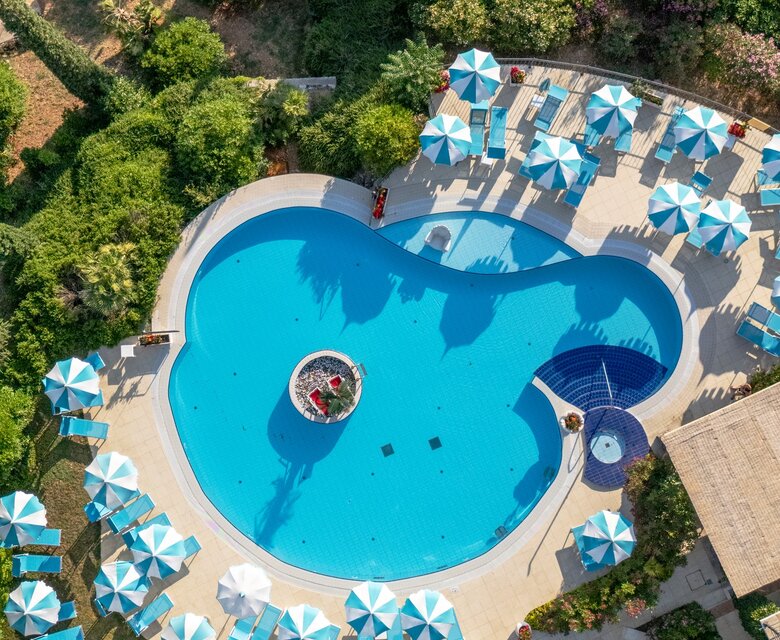 Ein Blick von oben auf einen einladenden Pool in Herzform, umgeben von grünen Gärten und zahlreichen blauen Sonnenschirmen. Die entspannte Atmosphäre lädt zum Schwimmen und Entspannen ein.