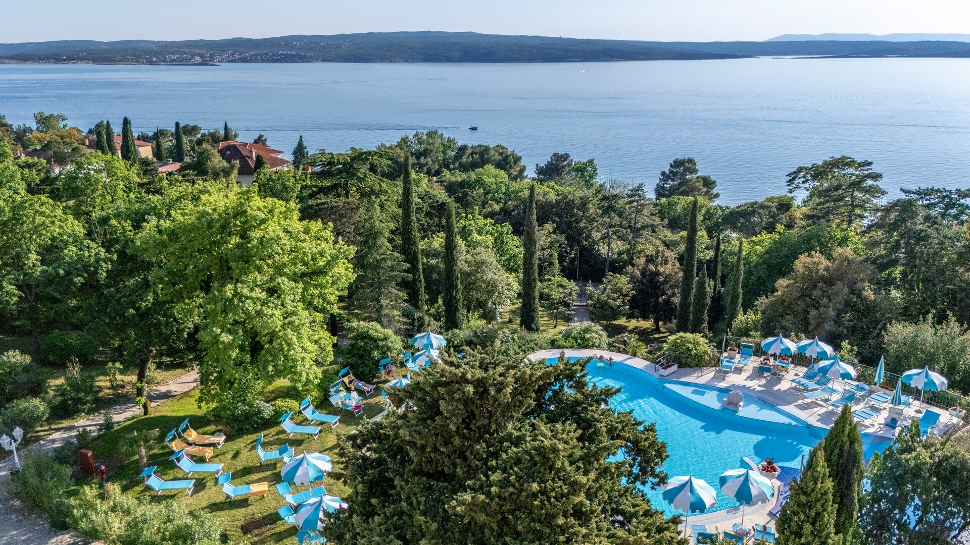 Blick auf einen einladenden Poolbereich umgeben von üppigem Grün und hohen Zypressen, mit Liegen und Sonnenschirmen. Im Hintergrund erstreckt sich ein ruhiger See und eine hügelige Landschaft.