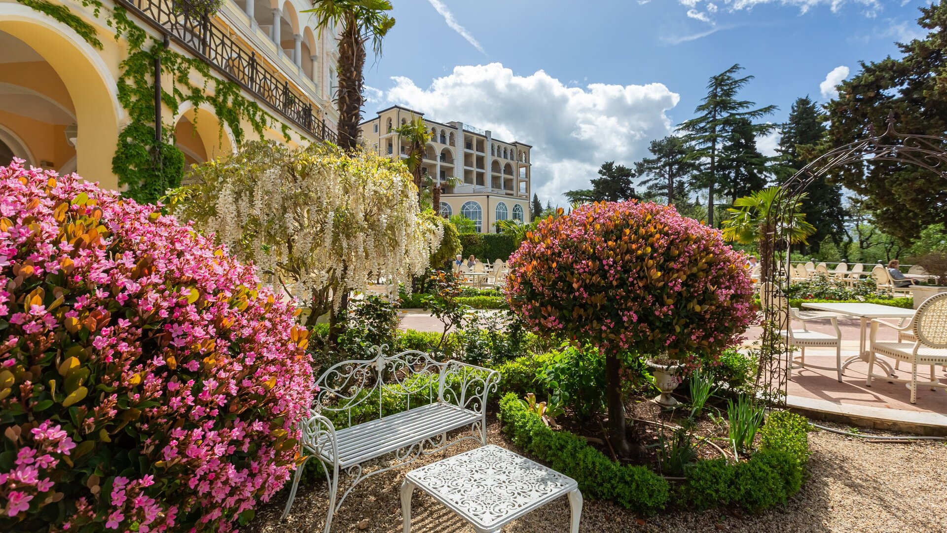 Ein blühender Garten mit bunten Blumen, darunter rosa und weiße Sträucher. Im Vordergrund steht eine weiße Bank und ein Tisch. Im Hintergrund ist ein elegantes Gebäude und blauer Himmel mit einigen Wolken zu sehen.