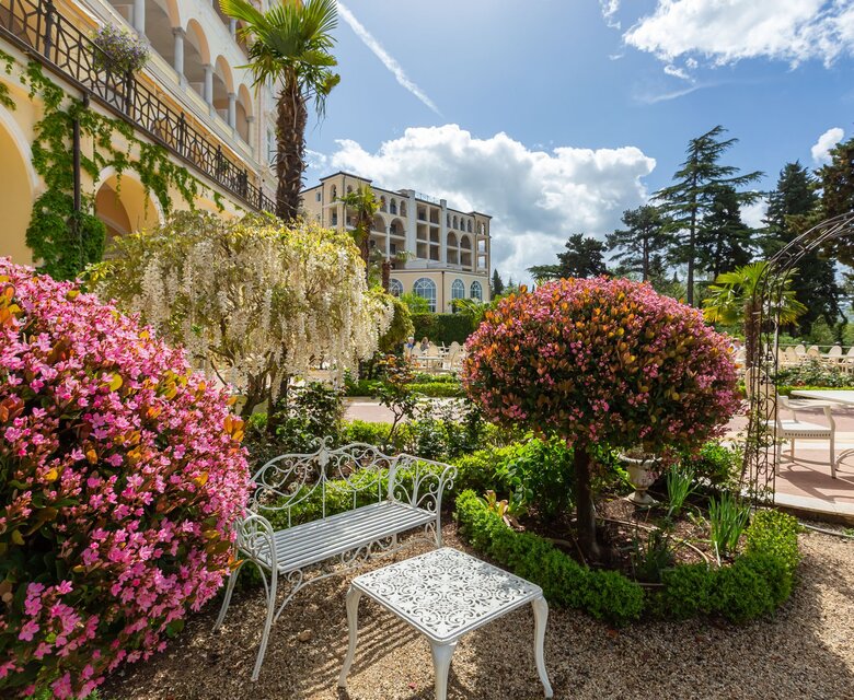 Ein blühender Garten mit bunten Blumen, darunter rosa und weiße Sträucher. Im Vordergrund steht eine weiße Bank und ein Tisch. Im Hintergrund ist ein elegantes Gebäude und blauer Himmel mit einigen Wolken zu sehen.