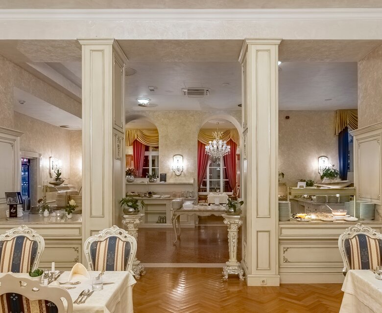Elegantes Restaurant mit stilvollen Tischen und Stühlen. Im Hintergrund sind Buffets und eine geschmackvolle Dekoration zu sehen. Die Wände sind in sanften Farben gehalten, und große Fenster lassen warmes Licht herein. Ein Kronleuchter sorgt für eine festliche Atmosphäre.