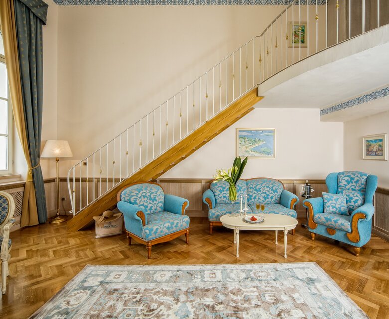 Heller Wohnbereich mit einer Treppe im Hintergrund. Zwei blaue Sessel und ein kleiner Tisch stehen auf einem eleganten Holzboden. Ein Teppich und eine Stehlampe ergänzen die gemütliche Atmosphäre. Große Fenster lassen viel Tageslicht herein.