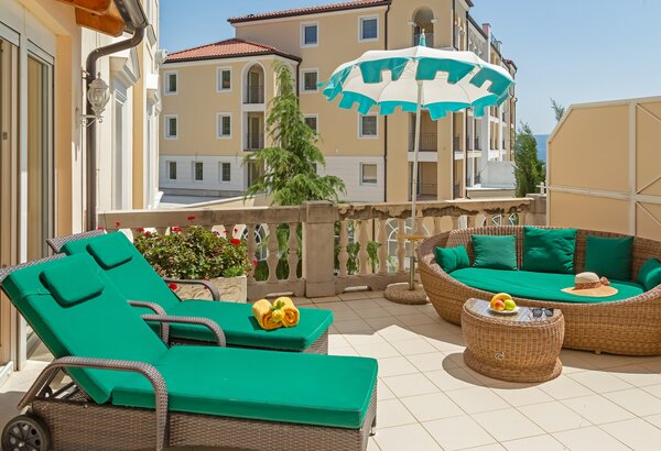 Eine sonnige Terrasse mit zwei grünen Liegen, einem runden Sofa und einem Tisch. Ein großer Sonnenschirm bietet Schatten. Im Hintergrund sind hellgelbe Gebäude und grüne Pflanzen sichtbar, die eine... Hotel mit Appartements für Familien, Kroatien - am Meer