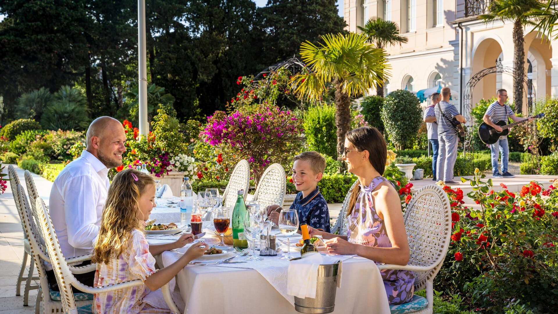 Eine Familie sitzt an einem schön gedeckten Tisch im Freien, umgeben von bunten Blumen und Palmen. Sie genießen ein Abendessen, während im Hintergrund Musiker spielen. Die Atmosphäre ist entspannt und einladend.