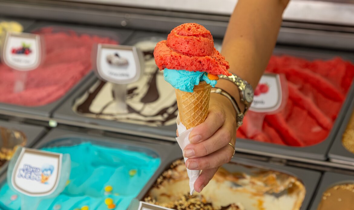 Eine Hand hält ein Waffeleis mit zwei Kugeln: einer roten und einer blauen. Im Hintergrund sind verschiedene Eissorten in Behältern sichtbar, die eine bunte Auswahl an Geschmäckern zeigen.