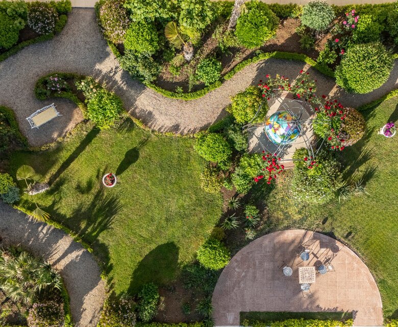 Luftaufnahme eines gepflegten Gartens mit geschwungenen Wegen, bunten Blumenbeeten und einer zentralen Sitzgruppe auf einer Terrasse. Grüne Sträucher und Bäume umrahmen die Szene, die eine einladende und ruhige Atmosphäre vermittelt.