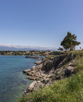Eine malerische Küstenlandschaft mit klarem, blauem Wasser, steinigen Ufern und sanften Hügeln. Im Hintergrund sind bunte Häuser und Bäume sichtbar, während der Himmel strahlend blau ist. Ideal für einen entspannten Aufenthalt am Meer.