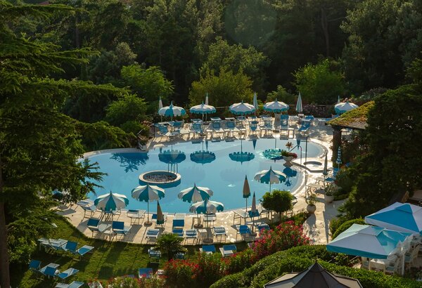 Ein einladender Poolbereich umgeben von üppigem Grün, mit blauen Liegen und Sonnenschirmen. Der glitzernde Pool spiegelt die Sonne wider, während Blumenbeete eine farbenfrohe Atmosphäre schaffen. Ideal für Entspannung und Erholung.