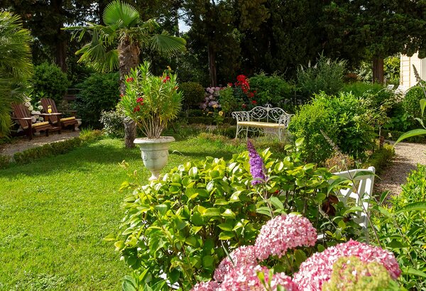 Ein einladender Garten mit üppigem Grün, bunten Blumen und einem kleinen Sitzbereich aus Holz. Im Hintergrund sind Sträucher und Bäume zu sehen, die eine ruhige Atmosphäre schaffen.