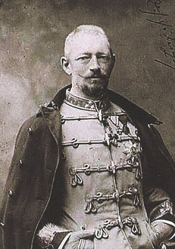 Ein historisches Porträt eines Mannes in einer militärischen Uniform mit dekorativen Knöpfen und Abzeichen. Er trägt einen Umhang und hat einen gepflegten Bart. Der Hintergrund ist unscharf und vermittelt eine nostalgische Atmosphäre.