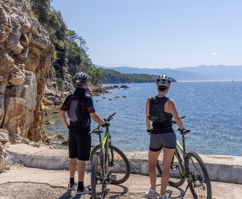 Zwei Radfahrer stehen am Ufer und blicken auf das ruhige Wasser. Die Küstenlandschaft ist von Felsen und Bäumen umgeben, während die Sonne am klaren Himmel scheint.