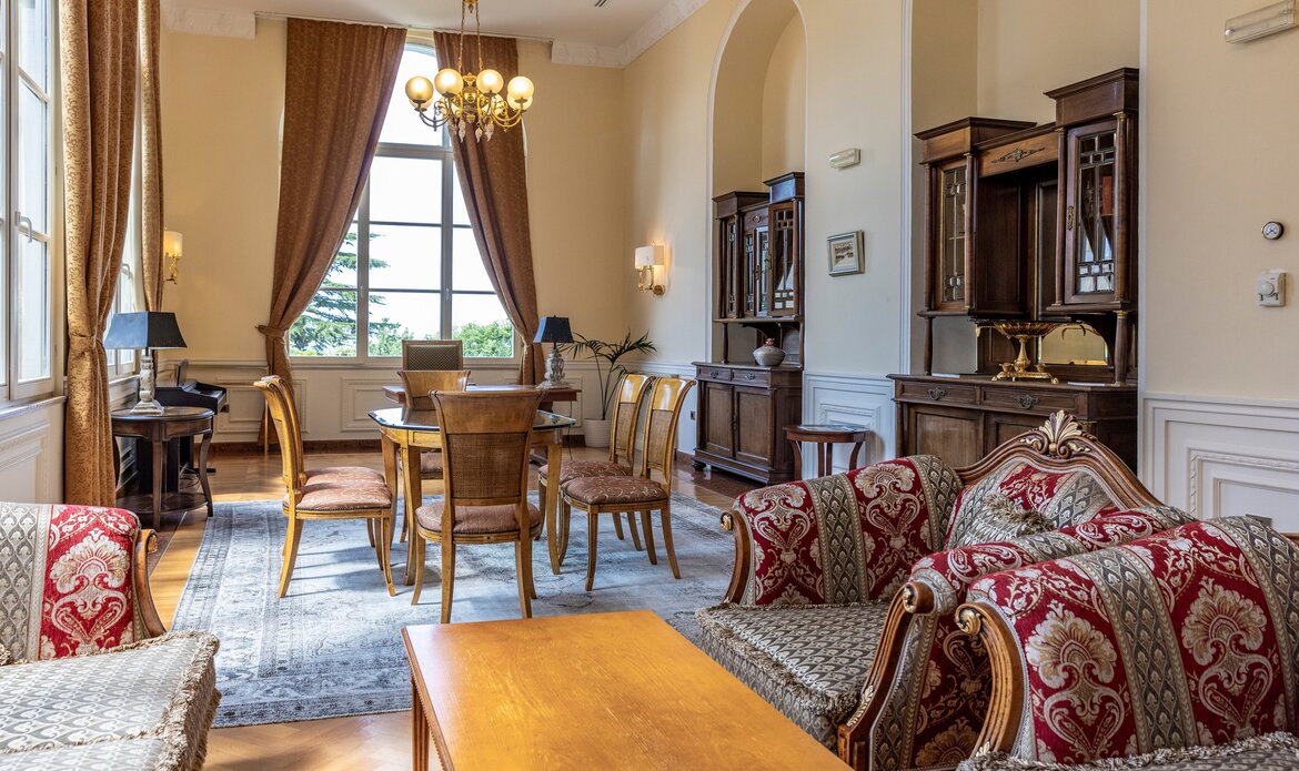 Elegantes Wohnzimmer mit einem großen Fenster, das viel Tageslicht hereinlässt. In der Mitte steht ein runder Esstisch mit Stühlen, umgeben von stilvollen Möbeln und einem Teppich. Gemütliche Sofas mit roten und goldenen Mustern laden zum Verweilen ein.