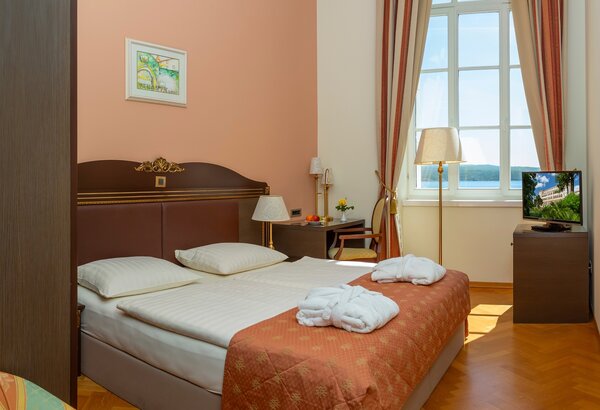 Ein gemütliches Hotelzimmer mit einem Doppelbett, zwei Kissen und frischen Handtüchern. Ein Fenster mit Vorhängen lässt Tageslicht herein, während ein Fernseher und ein kleiner Tisch mit Stuhl den ... Hotel mit Meerblick in der Kvarner Bucht - Doppelzimmer