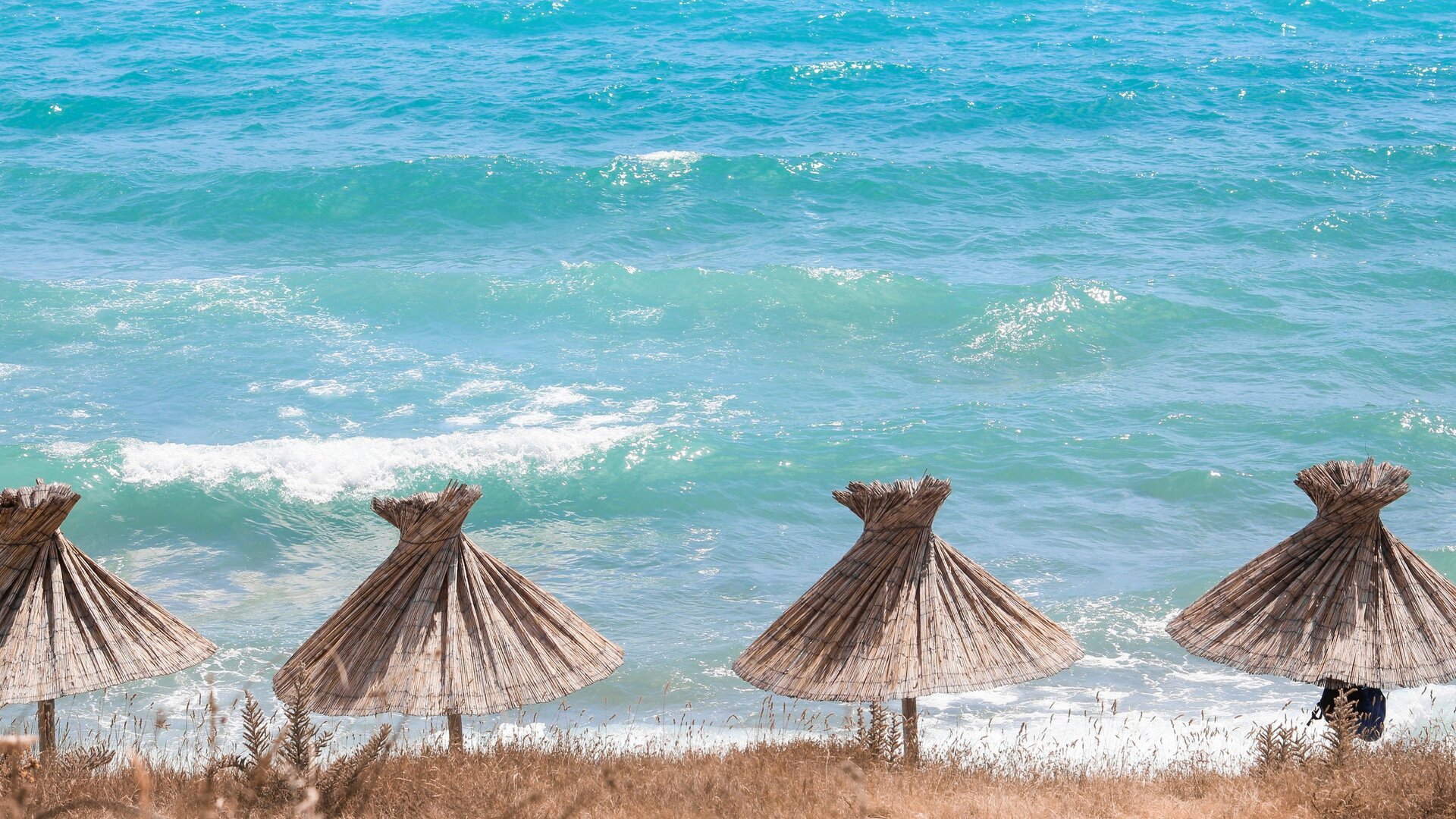 Strand mit mehreren Strohhütten im Vordergrund, die sanften Wellen des türkisfarbenen Meeres im Hintergrund. Die Atmosphäre ist entspannt und einladend, ideal für einen Urlaubstag am Strand.