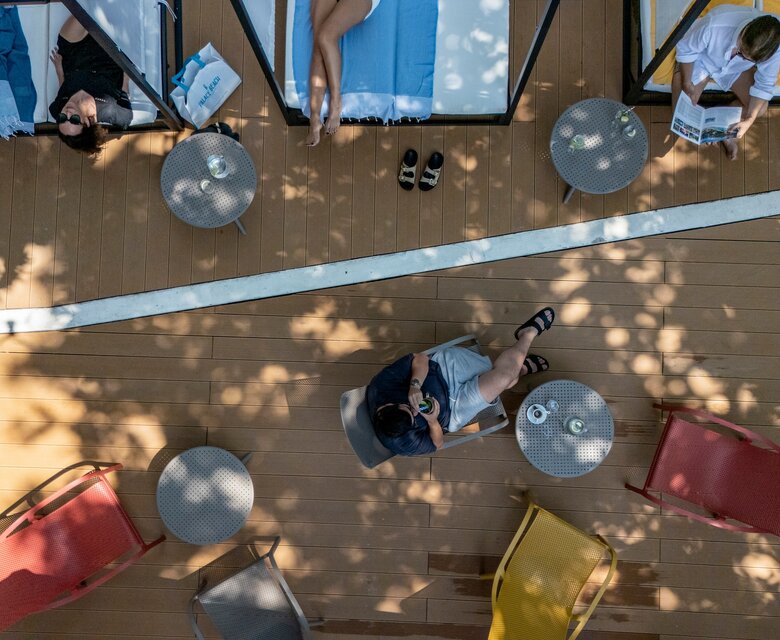 Eine Vogelperspektive zeigt eine entspannte Terrasse mit Liegen und Tischen. Einige Gäste lesen oder entspannen sich im Schatten von Bäumen, während bunte Stühle eine einladende Atmosphäre schaffen.