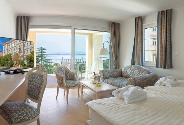 Helles Hotelzimmer mit zwei Betten, einem gemütlichen Sofa und einem Tisch. Ein Fernseher steht auf einem Schreibtisch. Große Fenster bieten einen Blick auf das Meer und einen Balkon mit Pflanzen. ... Hotel mit Appartements für Familien, Kroatien - am Meer
