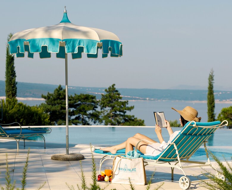 Eine Person entspannt auf einer Liege am Pool, liest ein Buch unter einem großen Sonnenschirm. Im Hintergrund erstreckt sich eine malerische Landschaft mit Wasser und Bäumen. Die Atmosphäre ist ruhig und einladend.