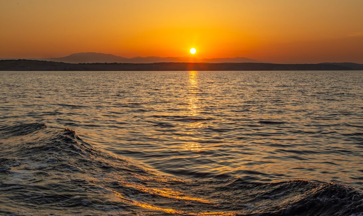 Sonnenuntergang über dem ruhigen Meer, mit sanften Wellen im Vordergrund und silhouettierten Bergen am Horizont. Die goldenen und orangefarbenen Farbtöne des Himmels spiegeln sich im Wasser wider.