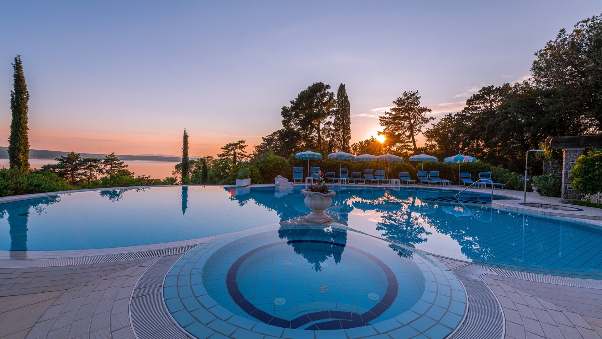 Ein ruhiger Poolbereich mit einem großen, glitzernden Pool, umgeben von grünen Bäumen und Liegen. Im Hintergrund ist ein Sonnenuntergang über dem Wasser zu sehen, der eine entspannte Atmosphäre schafft.