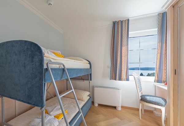 Ein helles Zimmer mit einem Etagenbett in Blau und weißen Bettwäsche. Neben dem Bett steht ein Stuhl mit einem gepolsterten Sitz. Ein Fenster mit Vorhängen bietet Blick auf die Umgebung. Der Boden ... Hotel mit Appartements für Familien, Kroatien - am Meer