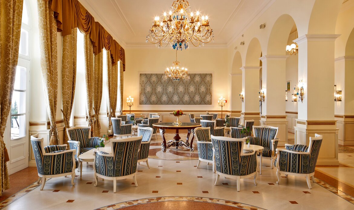 Eleganter Salon mit hohen Decken, großen Fenstern und goldenen Vorhängen. Mehrere stilvolle Sessel und Tische sind um einen zentralen Tisch gruppiert. Ein prächtiger Kronleuchter hängt von der Decke und sorgt für eine einladende Atmosphäre.
