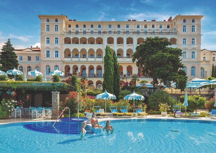 Ein luxuriöses Hotel mit einer eleganten Fassade und mehreren Etagen, umgeben von üppigen Gärten. Im Vordergrund ist ein blauer Pool, in dem einige Gäste schwimmen. Sonnenschirme und Liegen laden zum Entspannen ein.