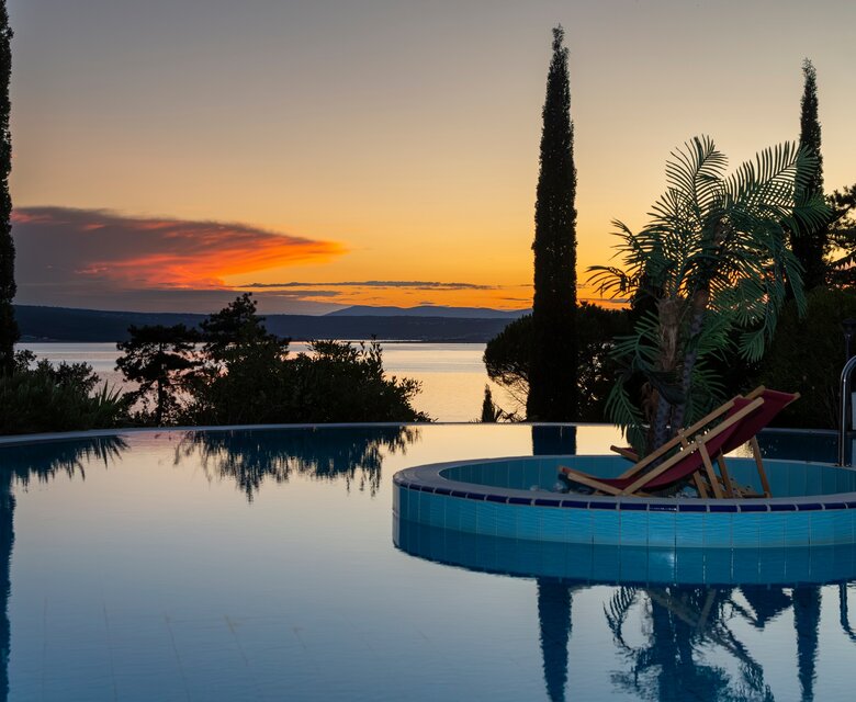 Ein ruhiger Pool mit einem Liegestuhl und Palmen, umgeben von Zypressen. Im Hintergrund leuchtet der Sonnenuntergang über dem Wasser, während die Farben des Himmels orange und lila strahlen.