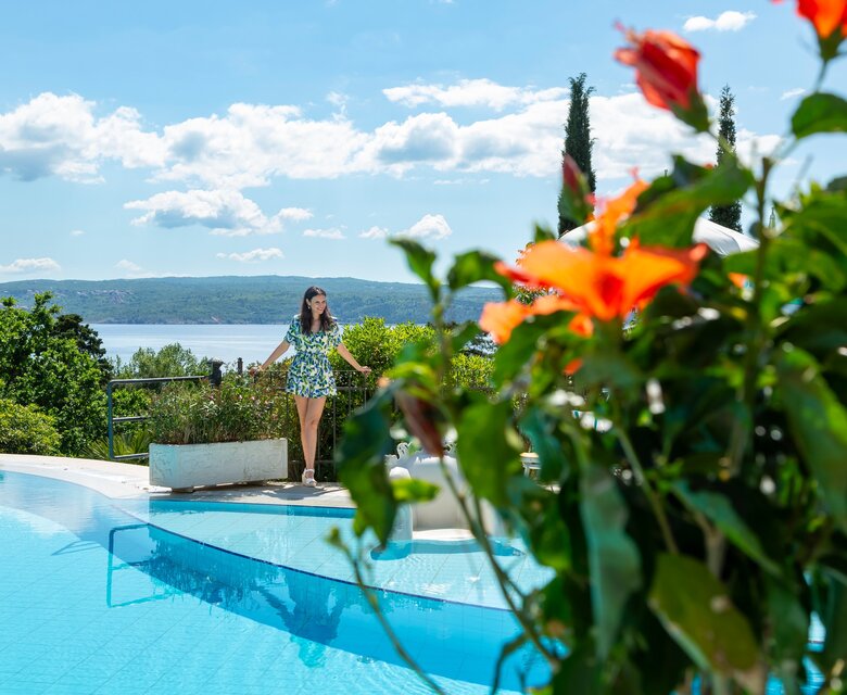Eine Frau steht am Rand eines glitzernden Pools, umgeben von blühenden Pflanzen. Im Hintergrund erstreckt sich ein schöner See unter einem klaren blauen Himmel mit einigen Wolken. Die Atmosphäre ist entspannt und einladend.