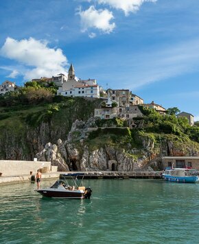 Ein malerischer Hafen mit einem kleinen Boot im Vordergrund, umgeben von klarem Wasser. Im Hintergrund erhebt sich eine steile Klippe mit charmanten Häusern und einer Kirche, unter einem strahlend blauen Himmel mit wenigen Wolken.