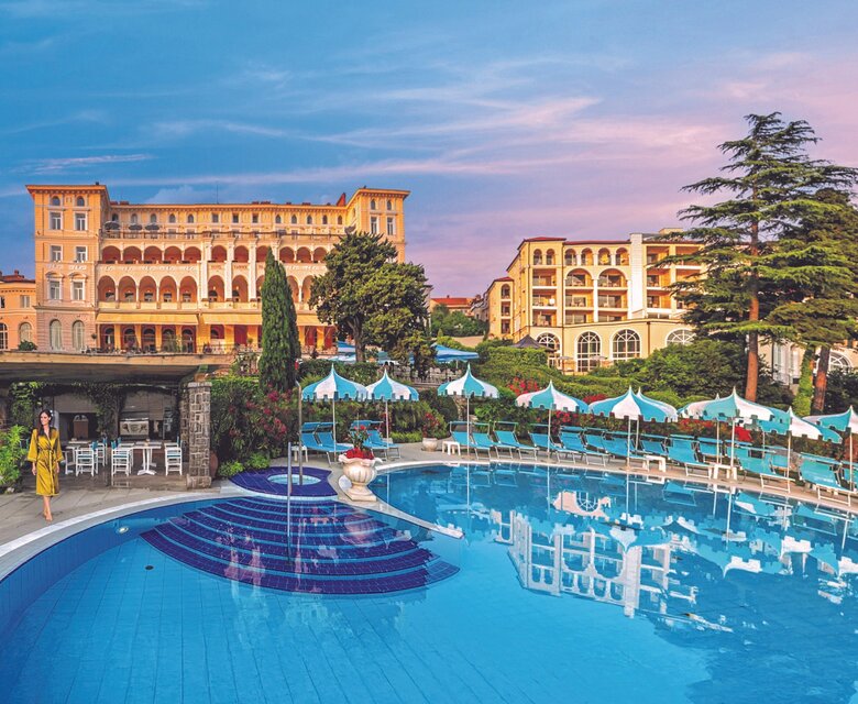 Ein elegantes Hotel mit einer großen, blauen Poollandschaft im Vordergrund. Umgeben von üppigem Grün und Bäumen, strahlt die Szenerie eine entspannte Atmosphäre aus. Der Himmel ist in sanften Pastellfarben gefärbt, was auf einen schönen Abend hindeutet.