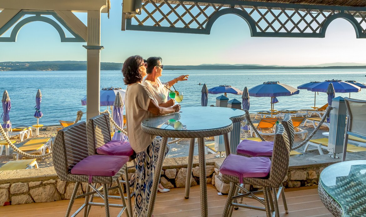 Zwei Frauen sitzen an einem Tisch in einer Strandbar mit Blick auf das Meer. Im Hintergrund sind Liegen und Sonnenschirme zu sehen. Die Atmosphäre ist entspannt und einladend, ideal für einen schönen Tag am Strand.