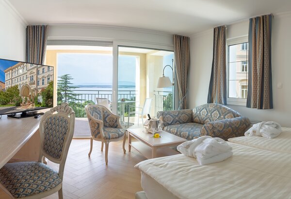 Helles Hotelzimmer mit zwei Betten, einem gemütlichen Sofa und einem Tisch. Große Fenster bieten einen Blick auf das Meer und einen Balkon. Ein Fernseher steht auf einem Schreibtisch. Die Einrichtung ist modern und einladend.