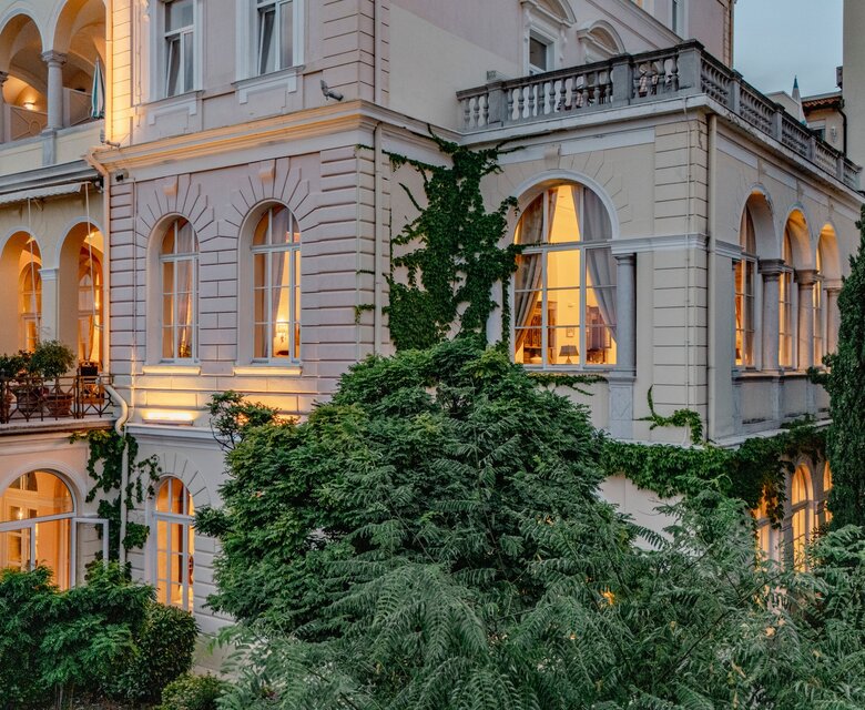 Eine elegante Hotelansicht mit großen Fenstern, durch die warmes Licht strahlt. Die Fassade ist mit Pflanzen bewachsen, umgeben von üppigem Grün. Die Architektur ist klassisch mit Bögen und Balkonen, die eine einladende Atmosphäre schaffen.