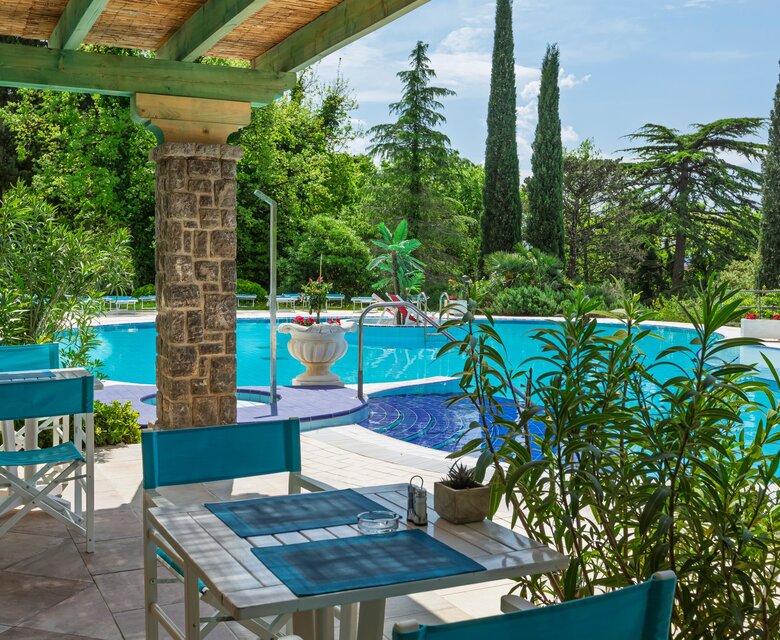Ein einladender Außenbereich mit weißen Tischen und blauen Stühlen, umgeben von üppigem Grün. Im Hintergrund befindet sich ein glitzernder Pool, umgeben von Palmen und Zypressen, unter einem strahlend blauen Himmel.