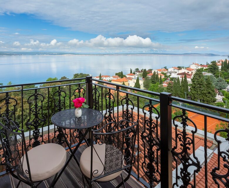 Blick von einem Balkon mit einem kleinen Tisch und zwei Stühlen, dekoriert mit einer Blume. Im Hintergrund erstreckt sich der ruhige See und eine malerische Stadt mit roten Dächern und grünen Bäumen unter einem bewölkten Himmel.