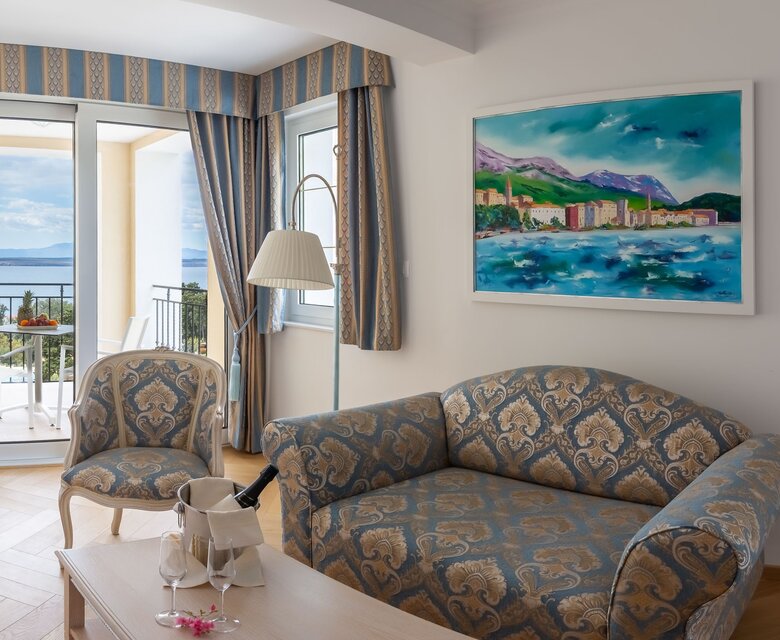 Helles Hotelzimmer mit einem gemütlichen Sofa und einem Sessel. Große Fenster bieten einen Blick auf das Meer und eine Terrasse. An der Wand hängt ein farbenfrohes Gemälde. Der Raum strahlt eine entspannte Atmosphäre aus.