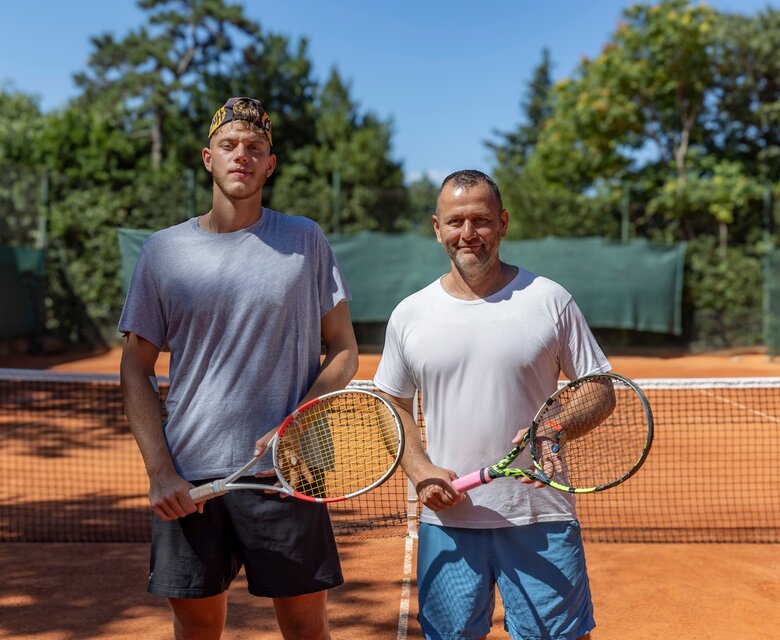Zwei Männer stehen auf einem Tennisplatz aus rotem Sand, jeder mit einem Tennisschläger in der Hand. Der jüngere Mann trägt ein graues T-Shirt und schwarze Shorts, während der ältere Mann ein weißes T-Shirt und blaue Shorts trägt. Im Hintergrund sind grüne Bäume und Tennisnetze sichtbar.