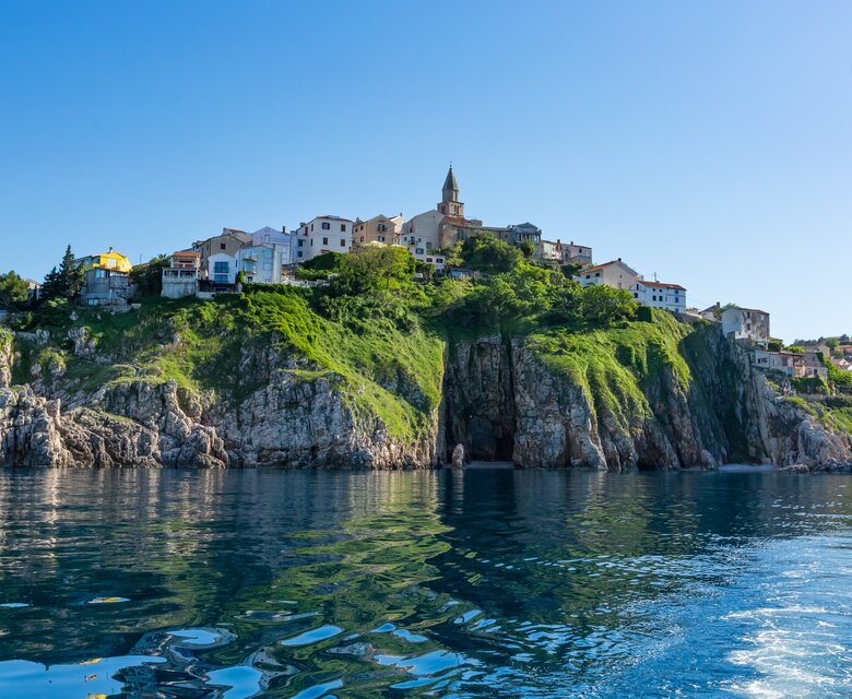 Eine malerische Küstenlandschaft mit einem Hügel, auf dem ein charmantes Dorf mit bunten Häusern und einer Kirche steht. Die Klippen sind von klarem, blauem Wasser umgeben, das die Sonne reflektiert.