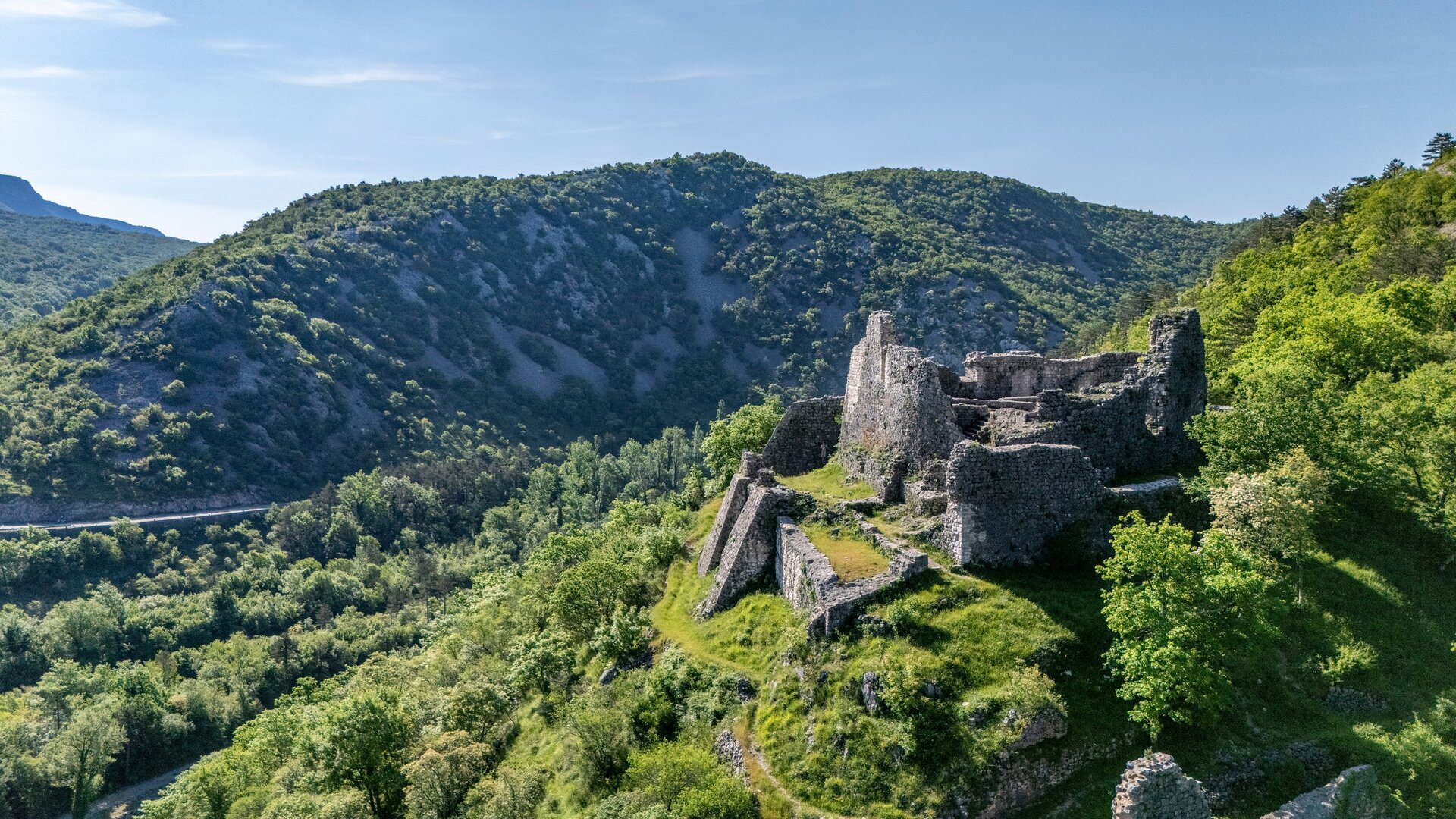 Eine beeindruckende Aussicht auf die Ruinen einer alten Burg, umgeben von üppigen grünen Wäldern und sanften Hügeln. Der Himmel ist klar und blau, was eine friedliche Atmosphäre schafft.