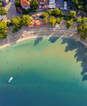 Luftaufnahme eines ruhigen Strandes mit Liegen, umgeben von Bäumen. Das klare Wasser zeigt sanfte Farbverläufe von Blau zu Grün. Im Hintergrund sind einige Gebäude und ein Boot auf dem Wasser zu sehen.